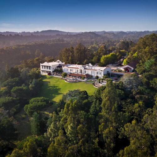 A sprawling estate with multiple white buildings nestled in a dense forest, perched on a hillside under a clear blue sky.