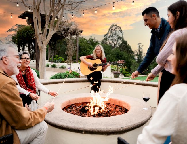 A woman plays guitar by a fire pit while a group sits and chats in a cozy outdoor gathering at sunset.