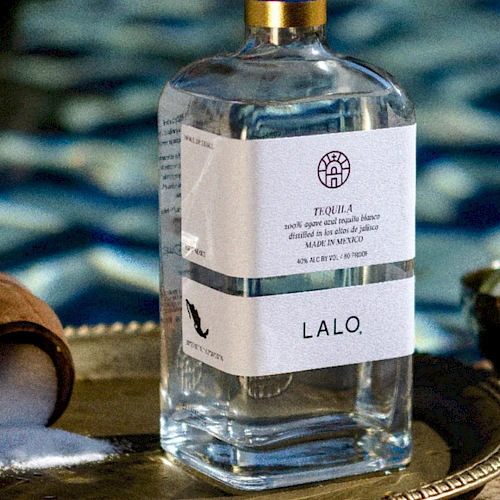 A bottle of LALO tequila sits on a decorative tray with a chocolate scoop, lime bowl, and a blurred pool background&mdash;simple luxury.