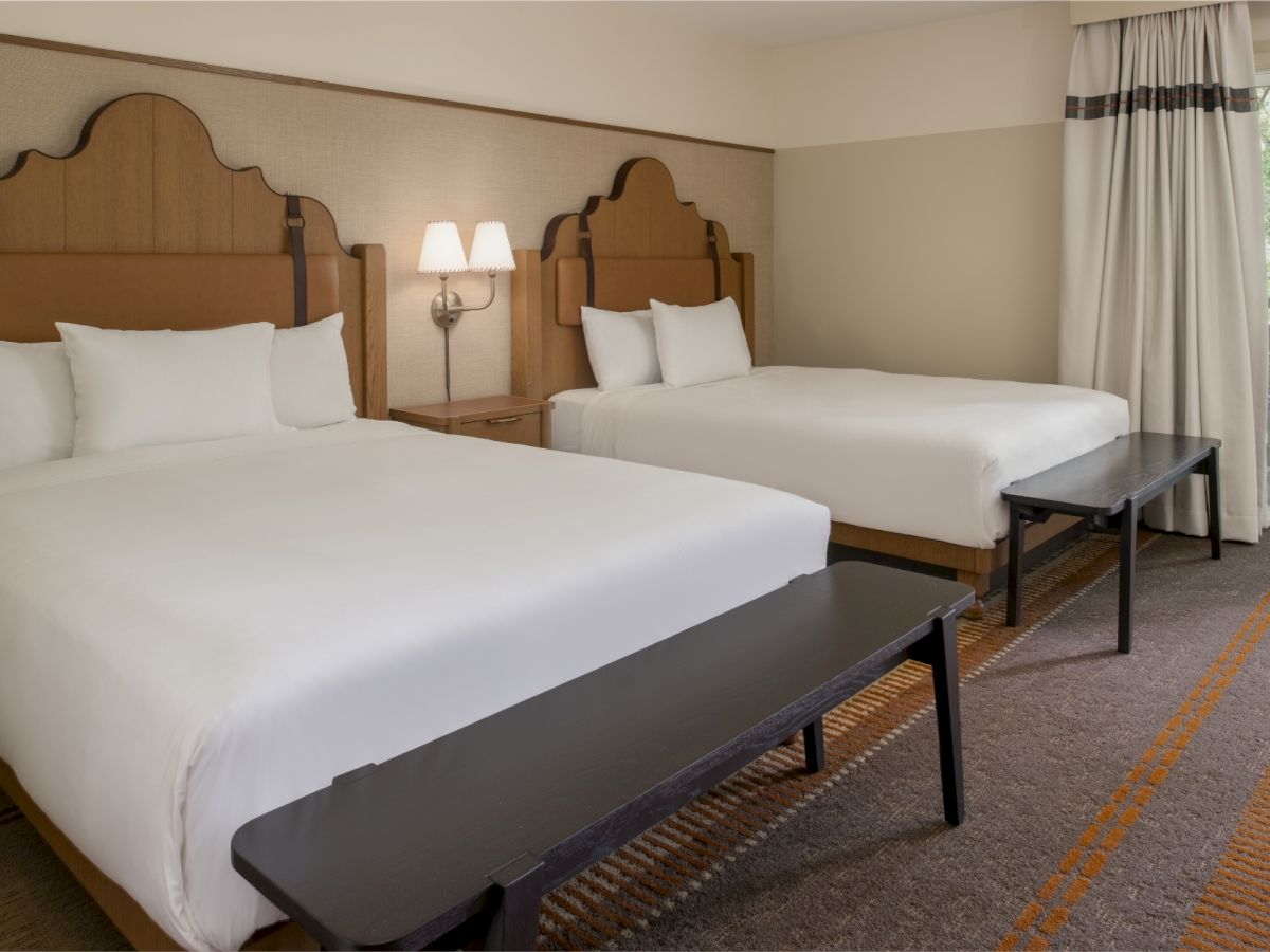 Two neatly made hotel beds with white linens, dark headboards, a bench at each foot, lamps on the wall, and a window with curtains.