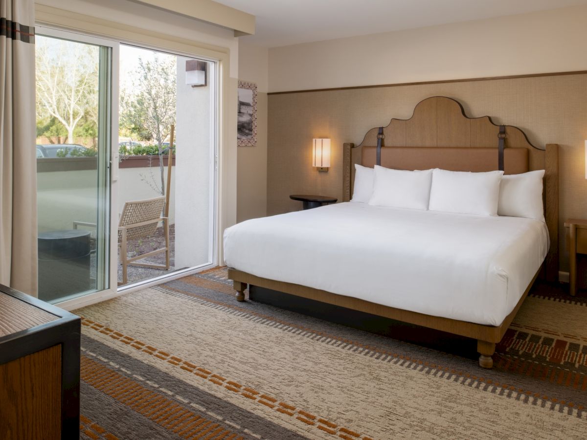 A modern hotel room with a king bed, two nightstands, a sliding glass door to a balcony, and warm lighting, creating a cozy retreat.