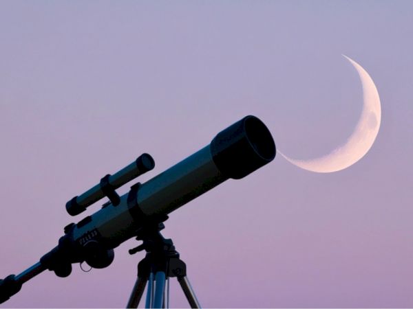 A blue telescope on a tripod aiming at a bright crescent moon against a twilight-purple sky.