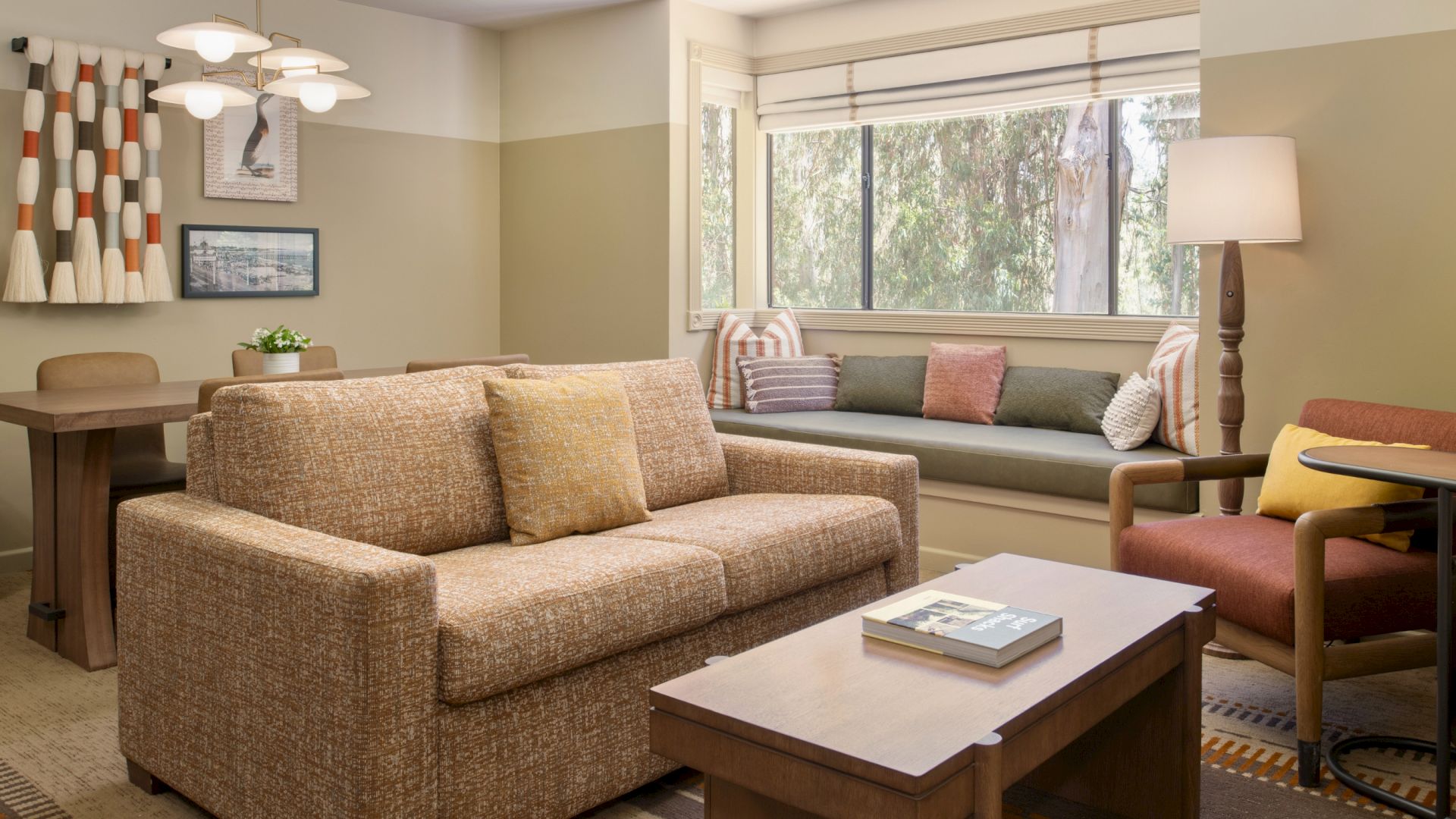 A bright living room with beige sectional sofa, colorful pillows, coffee table, lamps, and large window views.
