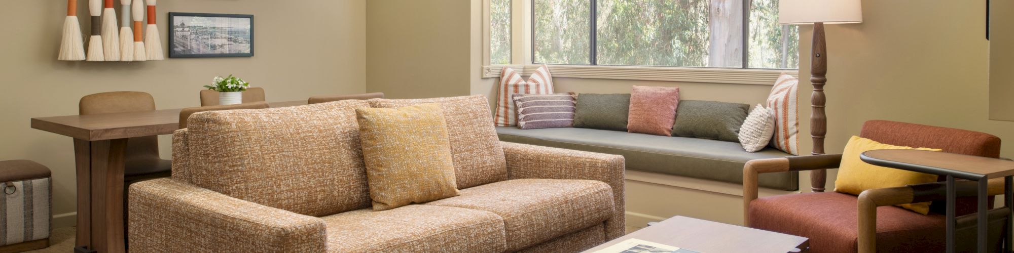 A bright living room with beige sectional sofa, colorful pillows, coffee table, lamps, and large window views.