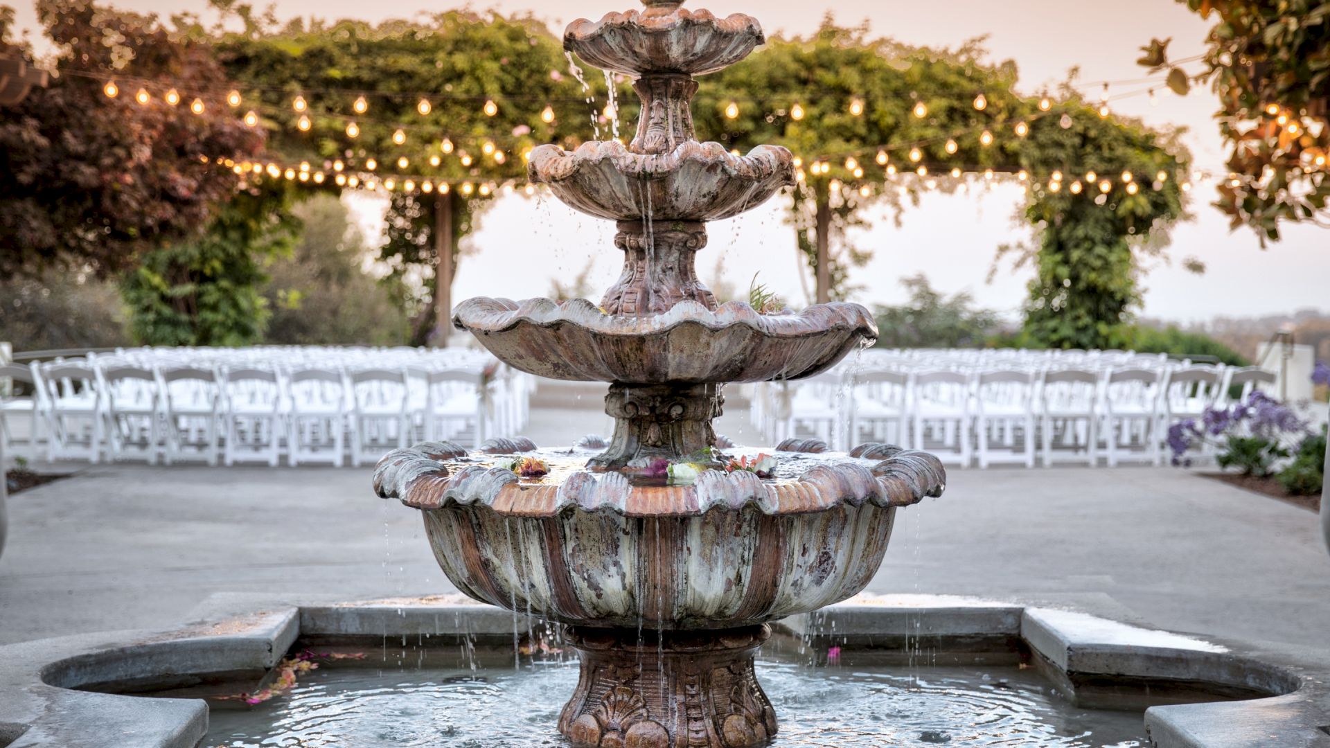 A decorative fountain is in the foreground, in front of outdoor seating and string lights under a canopy of greenery at dusk.