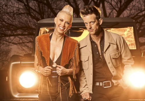 Two stylish people stand in front of a vintage jeep at dusk, smiling confidently as headlights glow behind them.