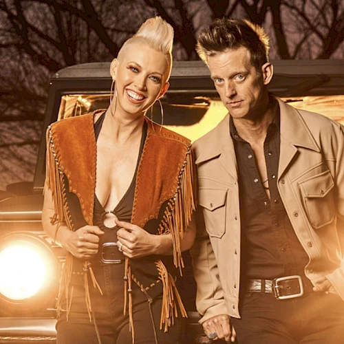 Two stylish people stand in front of a vintage jeep at dusk, smiling confidently as headlights glow behind them.