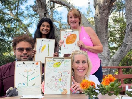 People outdoors holding colorful botanical artwork signs, posing with a fall-themed display under trees.