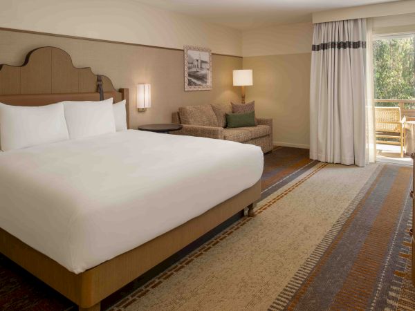 A hotel room with a large bed, nightstands, a sofa, and a balcony door, airy and well-lit, with neutral tones, cozy and inviting.