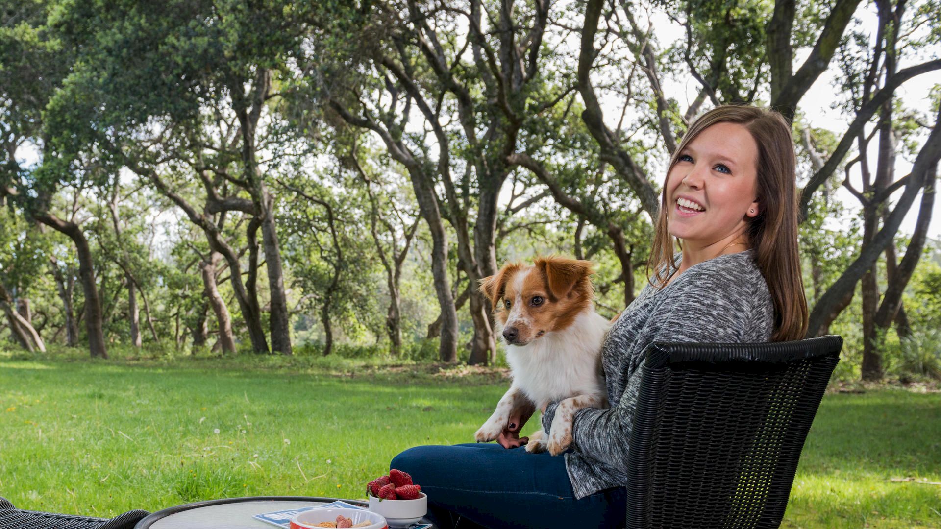 A woman sits outdoors in a chair with a dog on her lap, smiling and looking at the camera, surrounded by trees and a table with food.