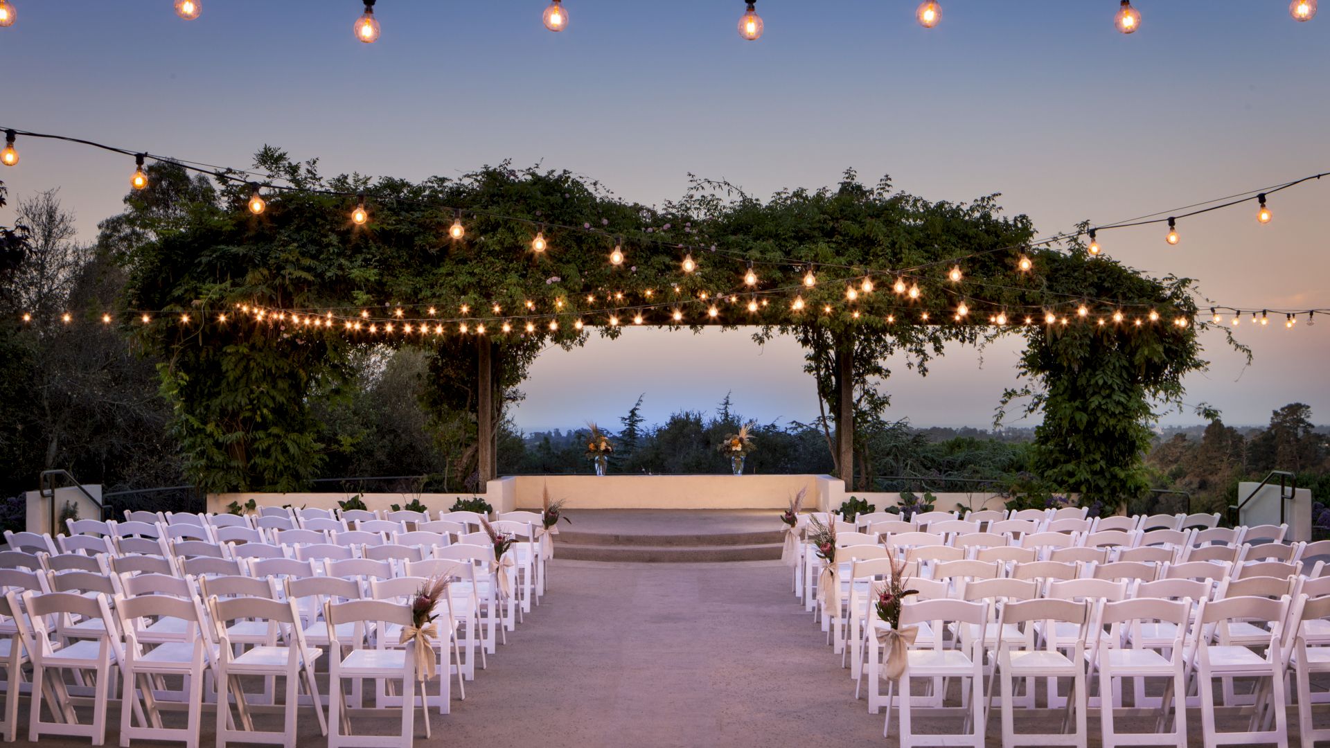 An outdoor wedding aisle at dusk with rows of white chairs, floral decor, and hanging string lights strung overhead.
