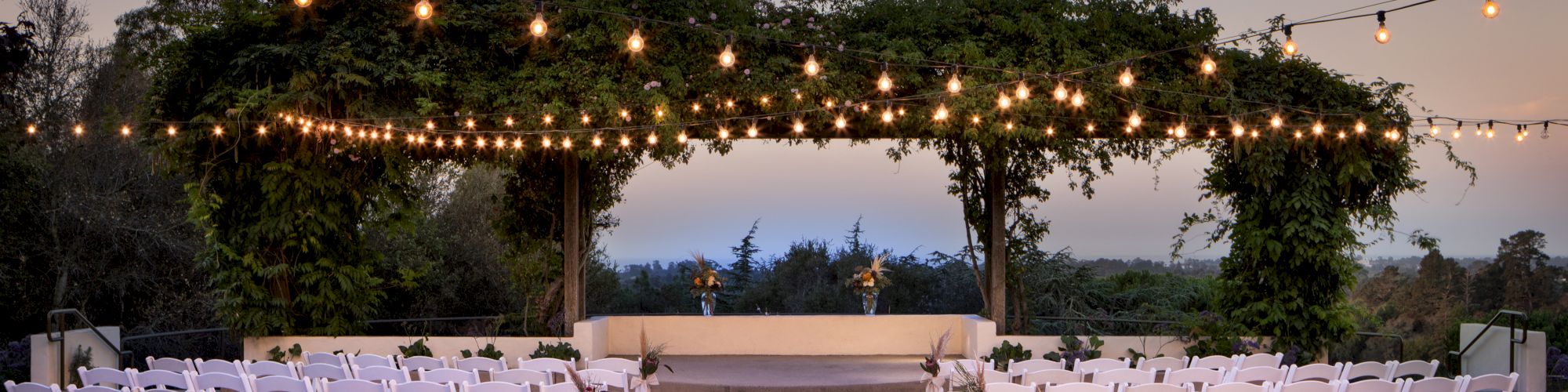 An outdoor wedding aisle at dusk with rows of white chairs, floral decor, and hanging string lights strung overhead.