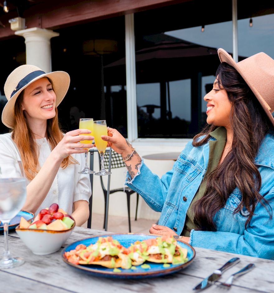Two women wearing hats share a sunny outdoor meal, clinking glasses over a plate of nachos and fruit on a terrace.