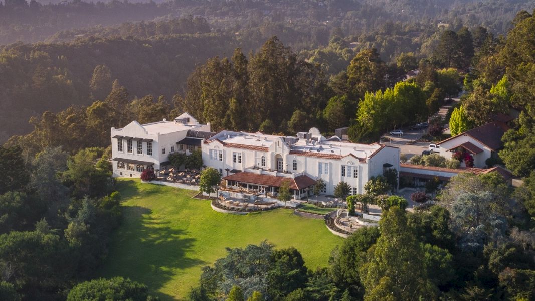 Aerial view of a large, luxurious estate surrounded by lush greenery and trees, with expansive grounds and multiple buildings.