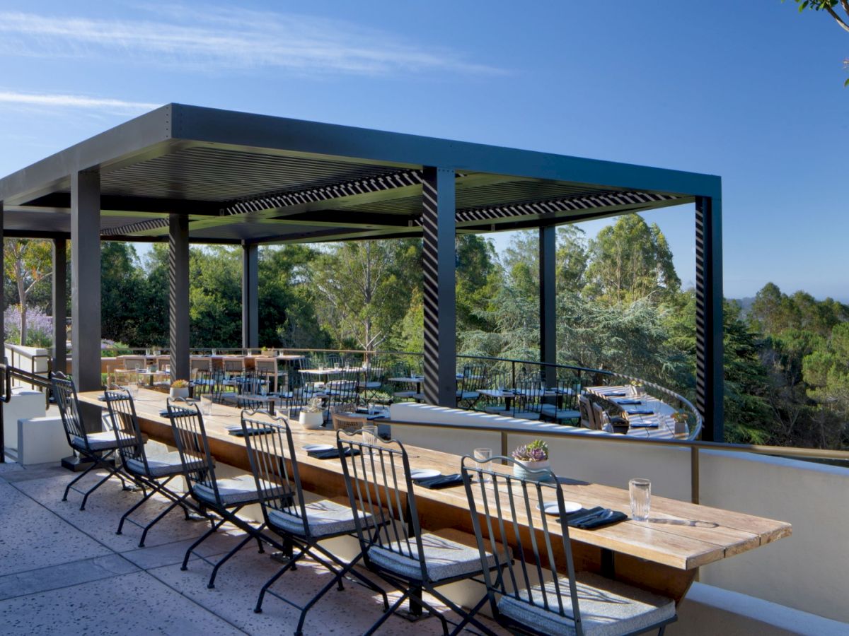 An outdoor event patio with long wooden tables and black chairs under a striped pergola, overlooking trees and a blue sky.