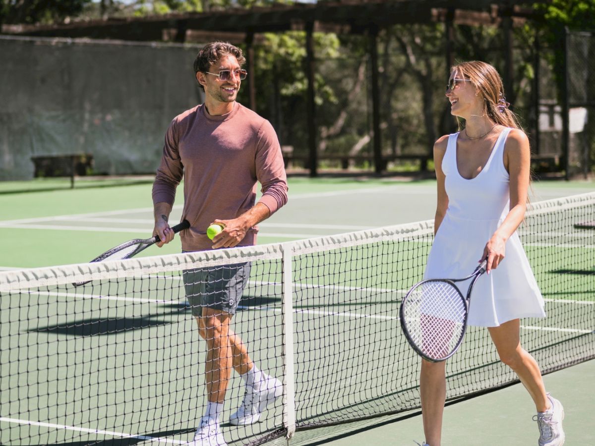 Two people walk a tennis court with rackets after play, chatting casually as they approach the net, wearing sporty outfits.