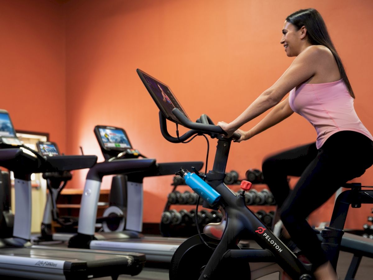 A woman rides a stationary bike in a gym with treadmills and weights, wearing a pink top and black leggings, focused on her workout.