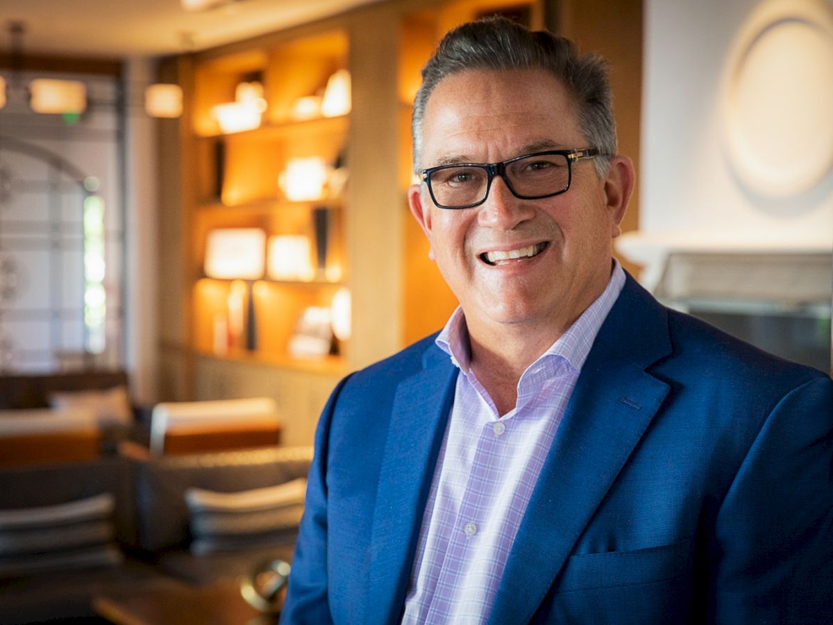 A smiling man in a blue blazer and glasses stands in a warmly lit, modern lounge with bookshelves in the background.