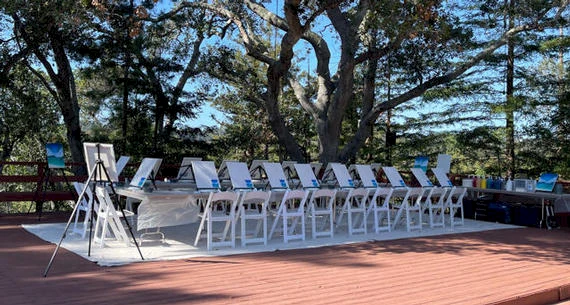 An outdoor painting setup with easels and chairs arranged in a semicircle on a deck surrounded by trees, under the shade of a large tree.