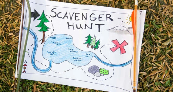 A colorful hand-drawn scavenger hunt map lies on grass, featuring a lake, trees, a red &ldquo;X,&rdquo; and an arrow compass pointing north.