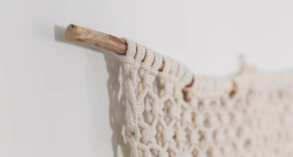 The image shows a close-up of a macram&eacute; wall hanging, with intricate knots, attached to a wooden dowel on a white wall.