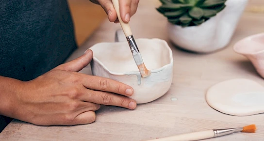 A person is painting a hand-crafted bowl with a brush, next to another ceramic piece and a succulent plant on a table.