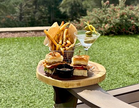 Outdoor table holds sliders, fries, dipping sauce, and a martini glass with olives in a backyard.