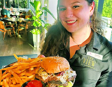 A smiling server in a restaurant stands by a tray with a cheeseburger and fries, with greenery-lit patio seating in the background.