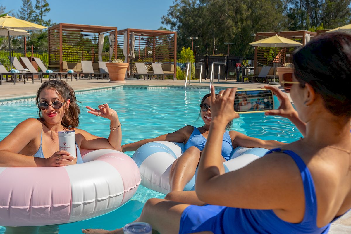 Friends float in an outdoor pool on inflatable rings, posing for a sunny selfie at a resort.