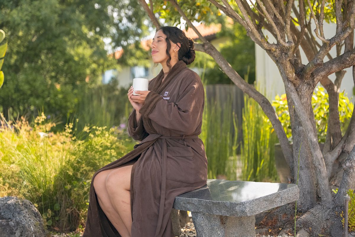 A woman in a brown robe relaxes outdoors, holding a white cup by a garden tree, soaking in the warm sun.