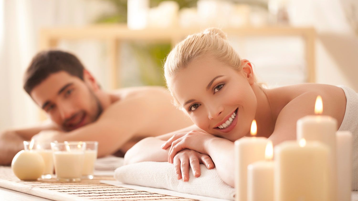 A man and a woman are lying down in a spa setting, surrounded by lit candles, appearing relaxed and content.