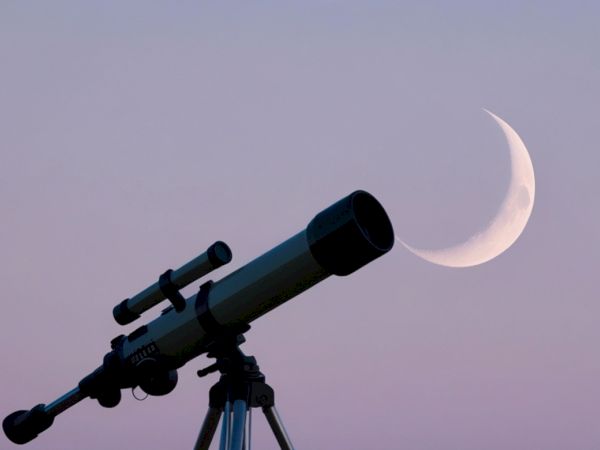 Silhouette of a telescope pointed at the crescent moon in the twilight sky.