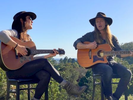 Two women in cowboy hats play acoustic guitars outdoors on stools against a sunny blue sky.
