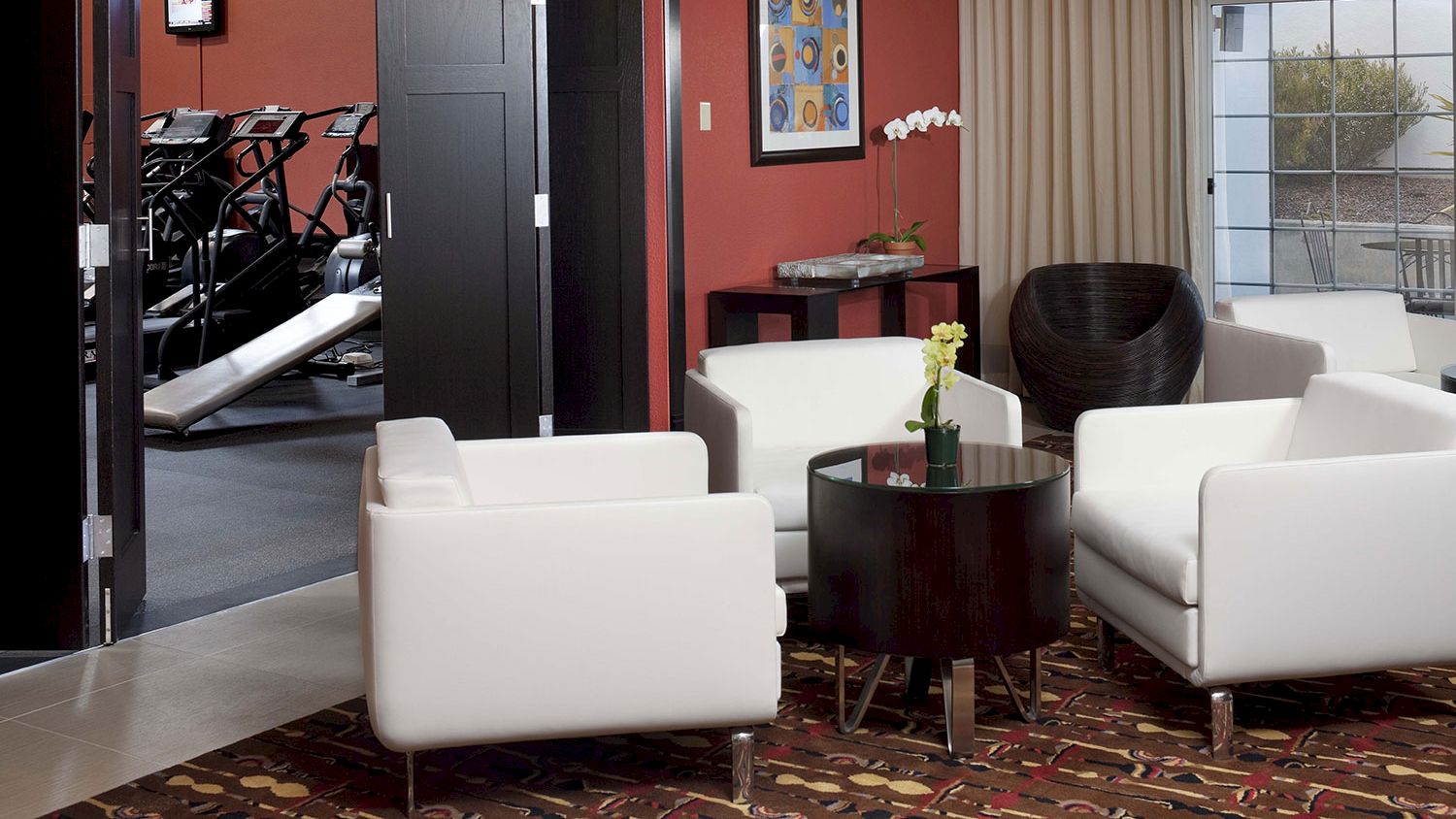 A modern waiting area with white chairs and a table, adjacent to a room with exercise equipment, decorated with a painting and a potted plant.
