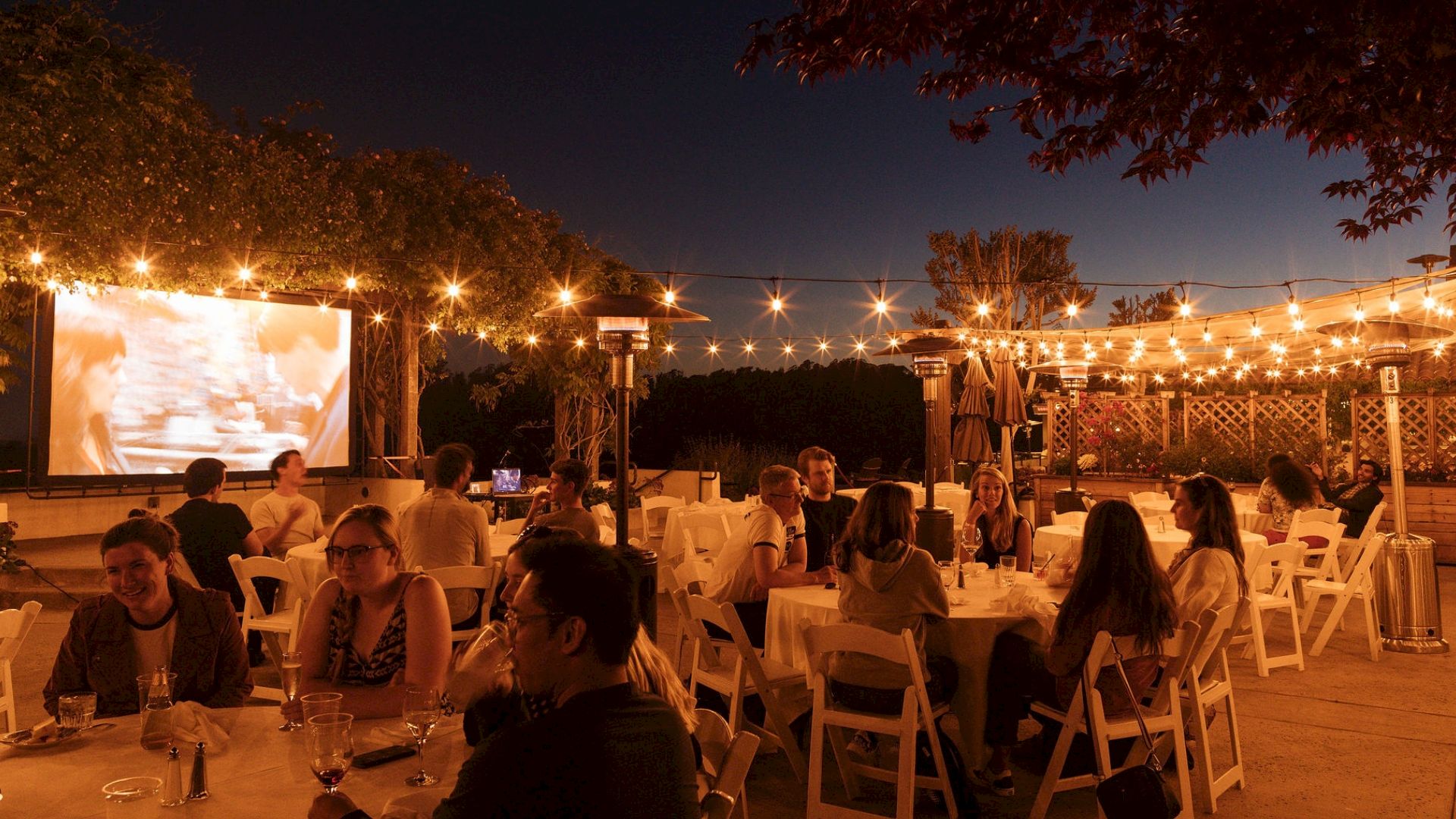 An outdoor dinner gathering with people seated at tables under warm string lights, with a movie projector on a screen.