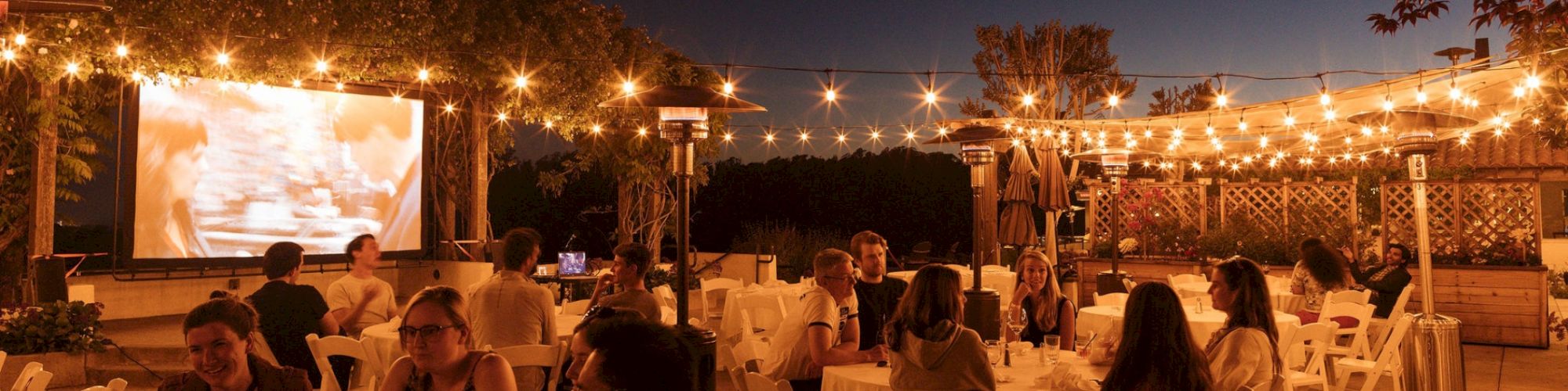 An outdoor dinner gathering with people seated at tables under warm string lights, with a movie projector on a screen.