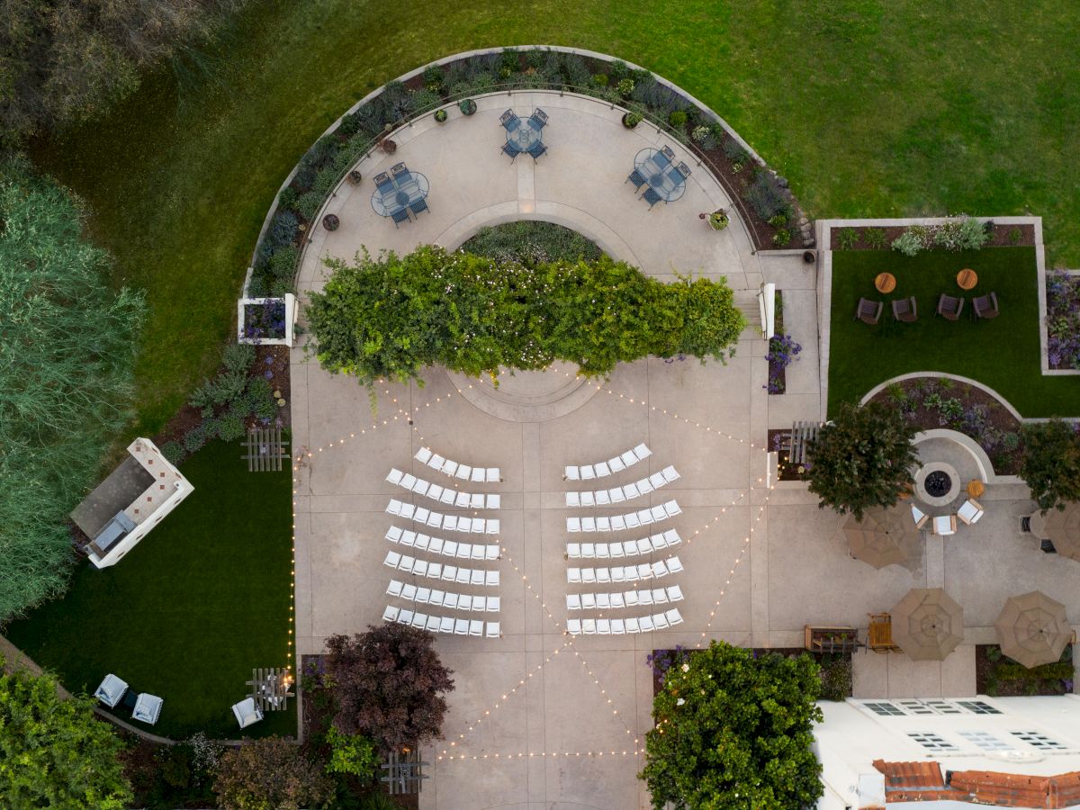 An aerial view of a landscaped courtyard set for a wedding: white chairs in rows, string lights, and round patio tables.