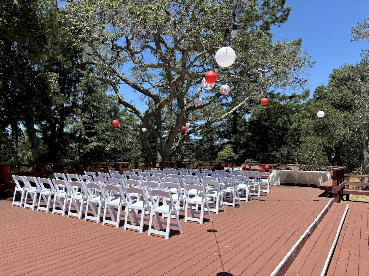 An outdoor ceremony deck with rows of white chairs and hanging red, white, and gold lanterns/ball lights from the trees.
