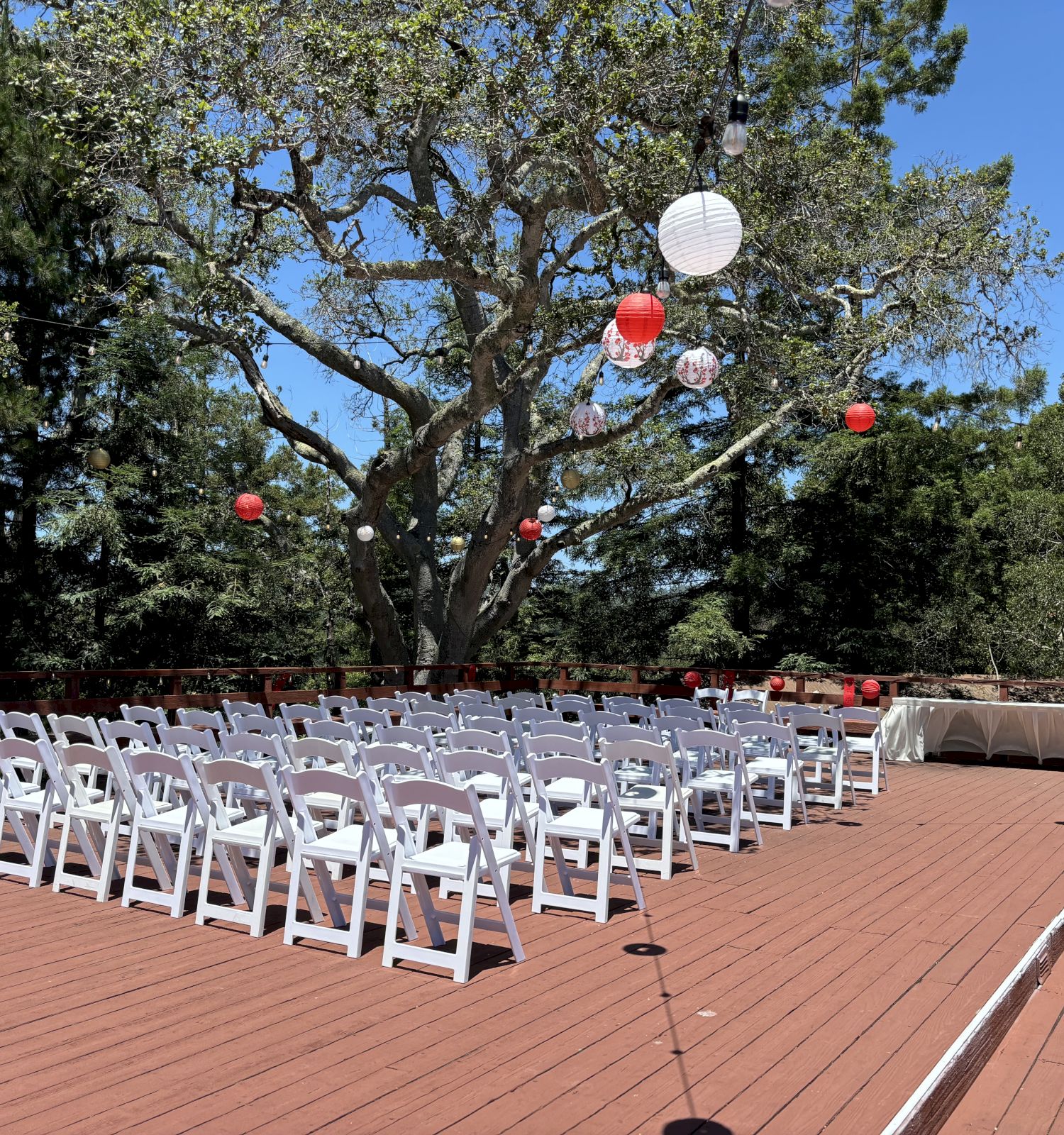 An outdoor ceremony deck with rows of white chairs and hanging red, white, and gold lanterns/ball lights from the trees.