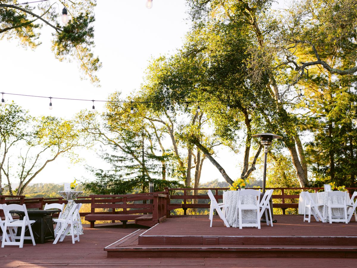 An outdoor patio with a row of white chairs, string lights, warm trees, and a fire pit for a gathering.