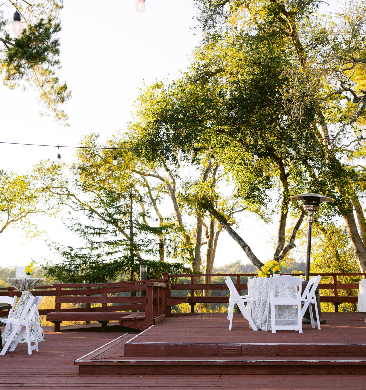 An outdoor patio with a row of white chairs, string lights, warm trees, and a fire pit for a gathering.