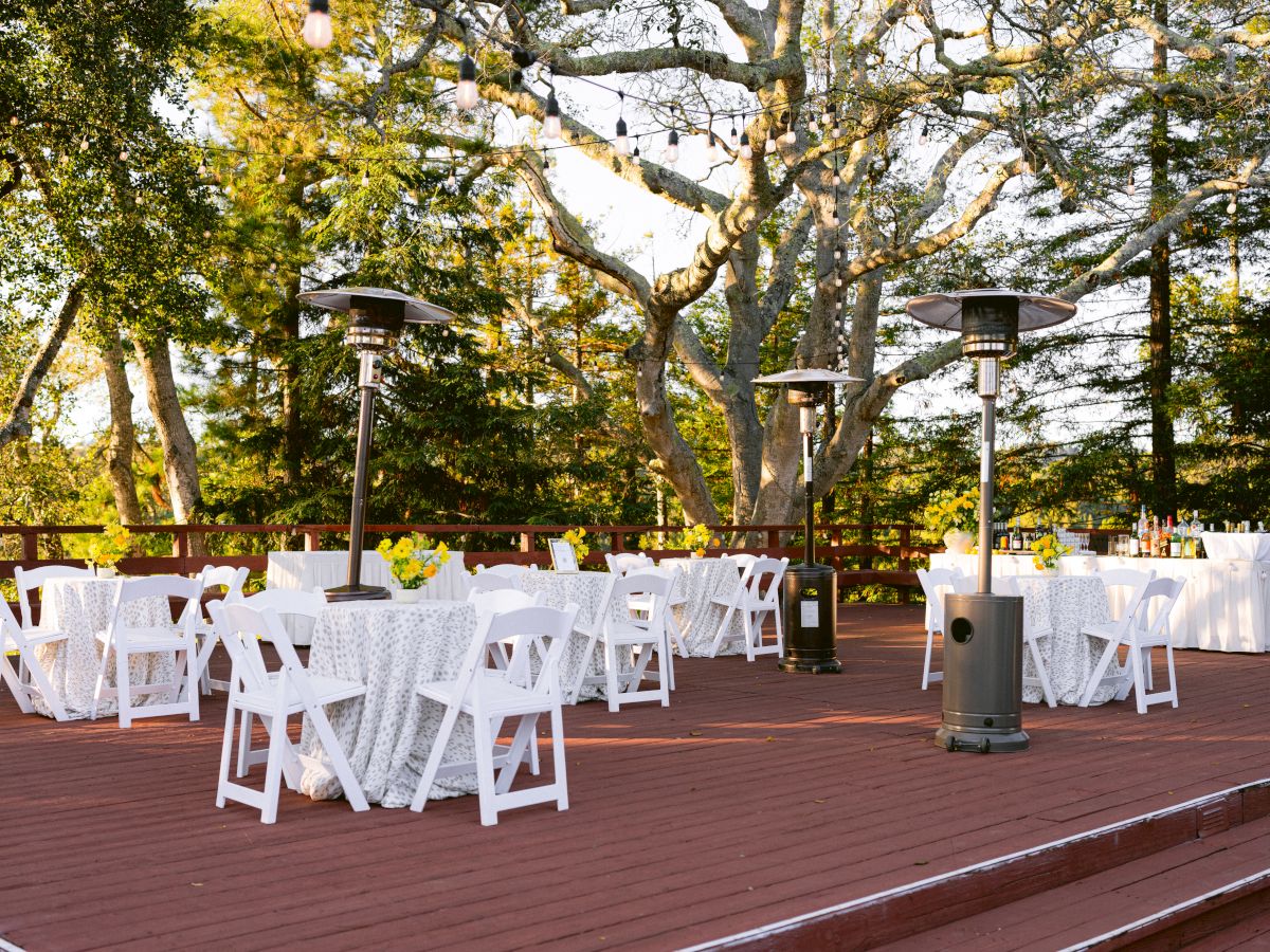 Outdoor wedding patio with white chairs, string lights, heaters, and a long decorated table under leafy trees.