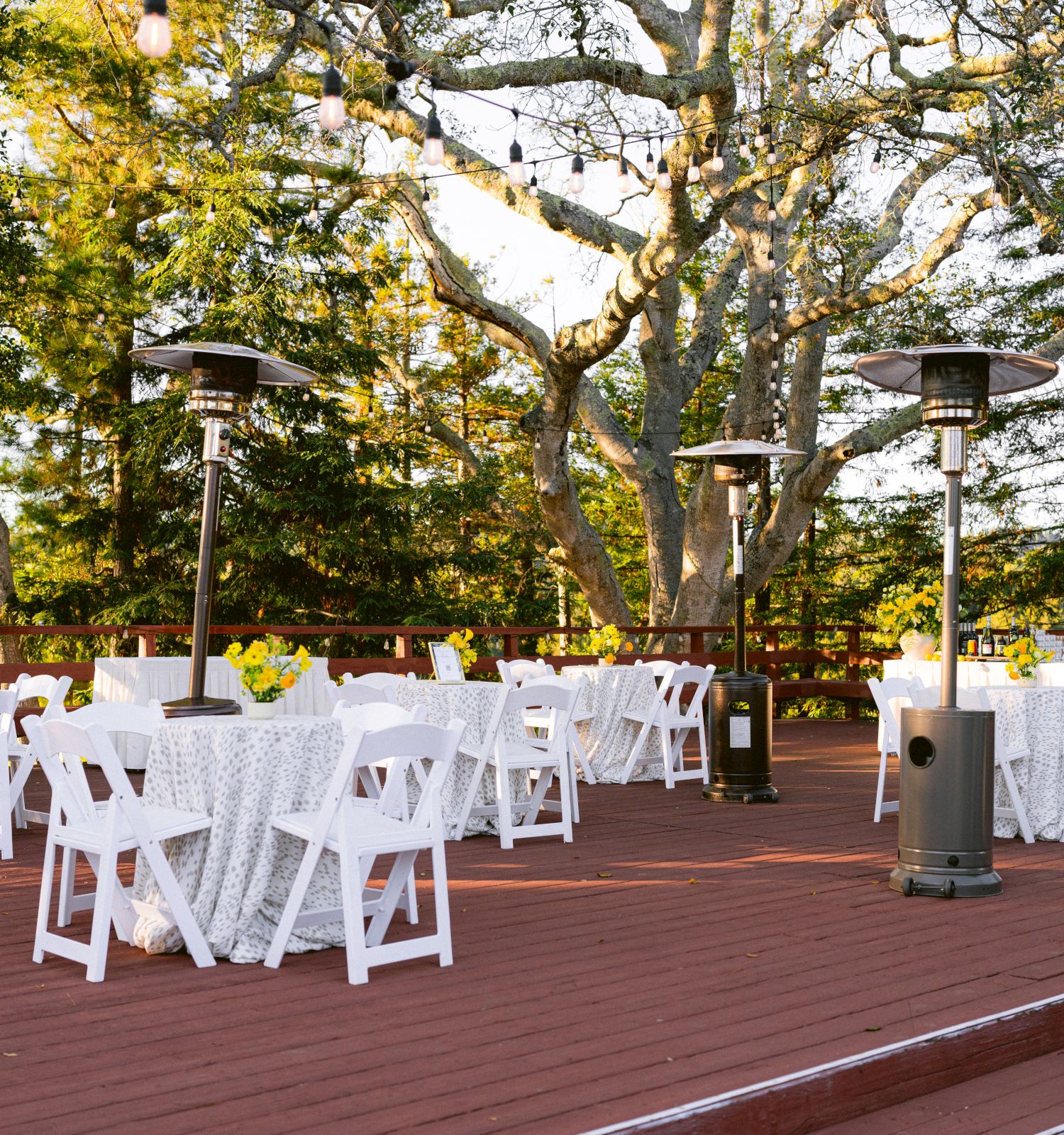 Outdoor wedding patio with white chairs, string lights, heaters, and a long decorated table under leafy trees.