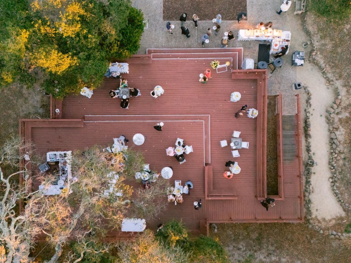 An overhead view of an outdoor event with guests seated at decorated tables and warm string lights, like a wedding or celebration.