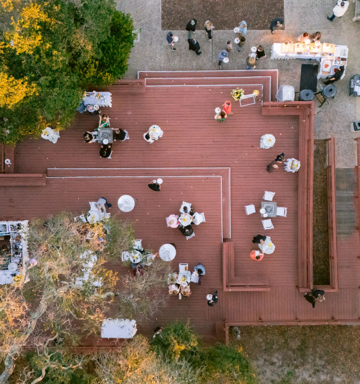 An overhead view of an outdoor event with guests seated at decorated tables and warm string lights, like a wedding or celebration.