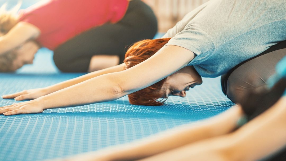 People in a yoga class practicing a child's pose on blue mats indoors, focused and stretching forward with their arms extended.