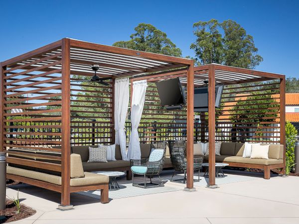 A modern outdoor seating area with two shaded pergolas featuring cushioned seats, a mounted TV, and small tables on a sunny day.