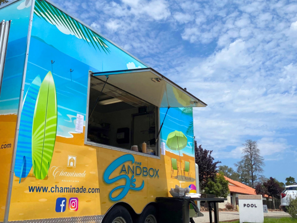 A vibrant food truck with beach-themed decor and social media icons is parked outdoors, near a pool sign and buildings, under a partly cloudy sky.
