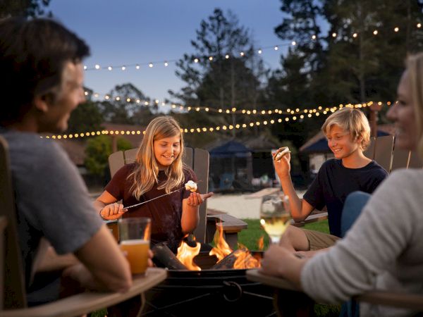 A group of four people sits around a fire pit, enjoying roasted marshmallows and drinks, with string lights overhead and trees in the background.