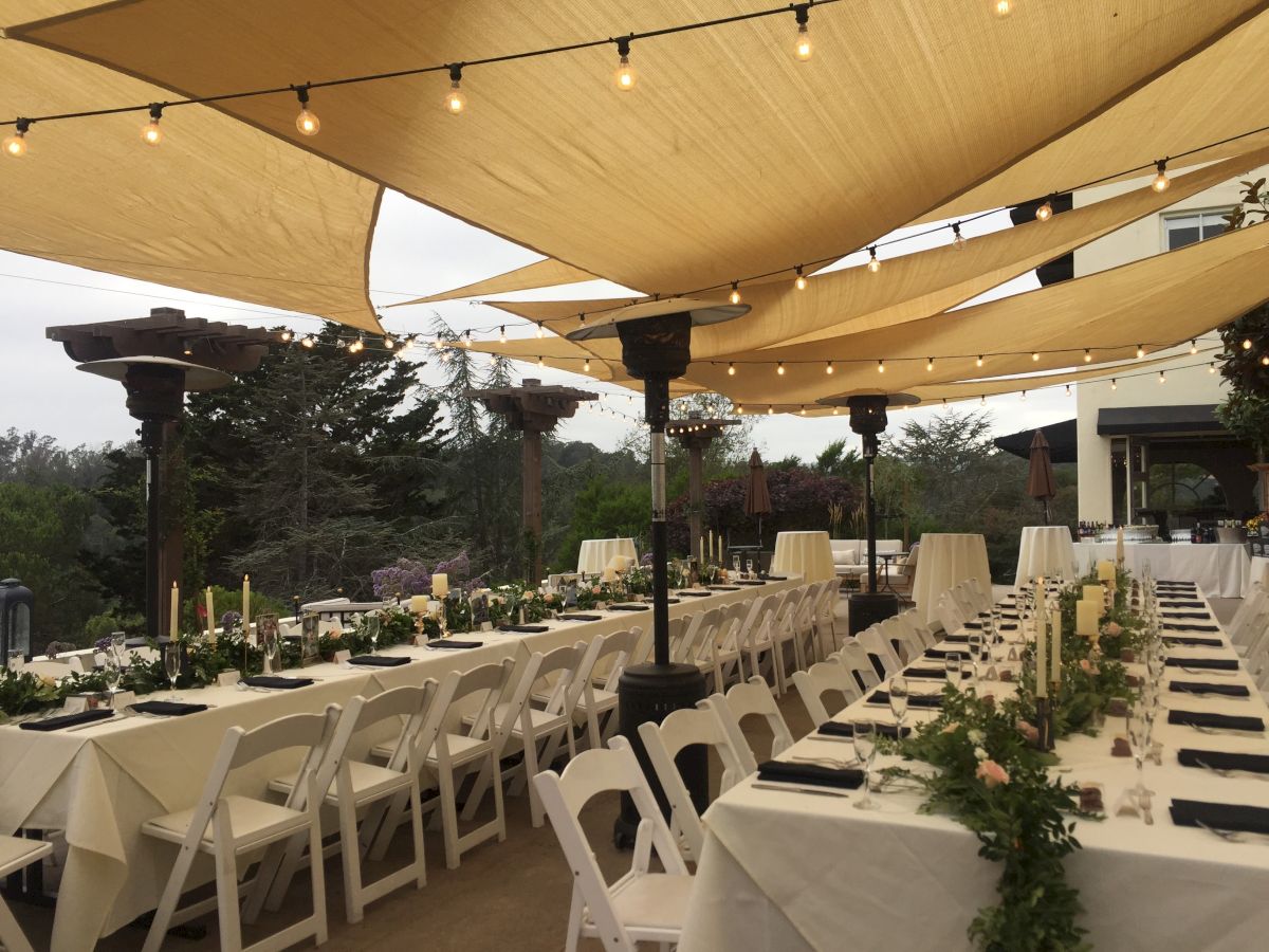 Outdoor wedding reception setup with white chairs, candlelit tables, and string lights under large beige canopy sails.